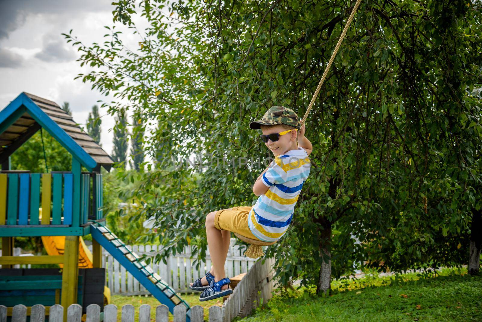 Happy little boy is having fun on a rope swing which he has found while having rest outside city. Active leisure time with children by Kobysh