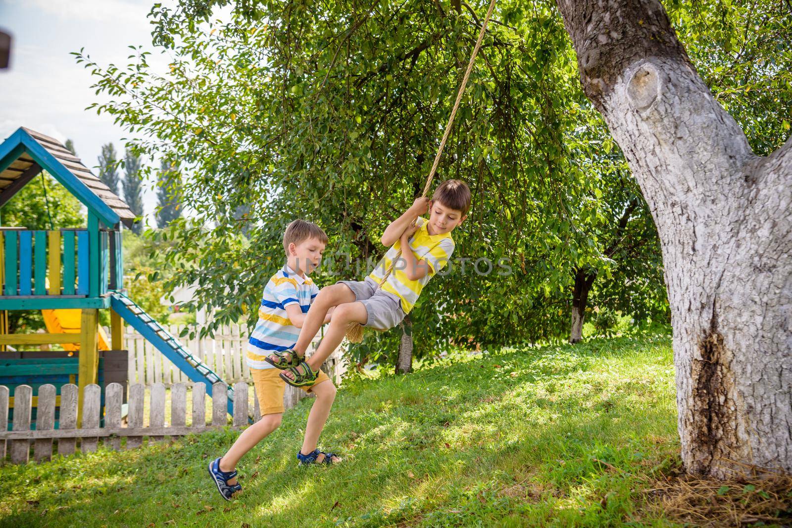 Two adorable happy little boys is having fun on a rope swing which he has found while having rest outside city. Active leisure time with children by Kobysh