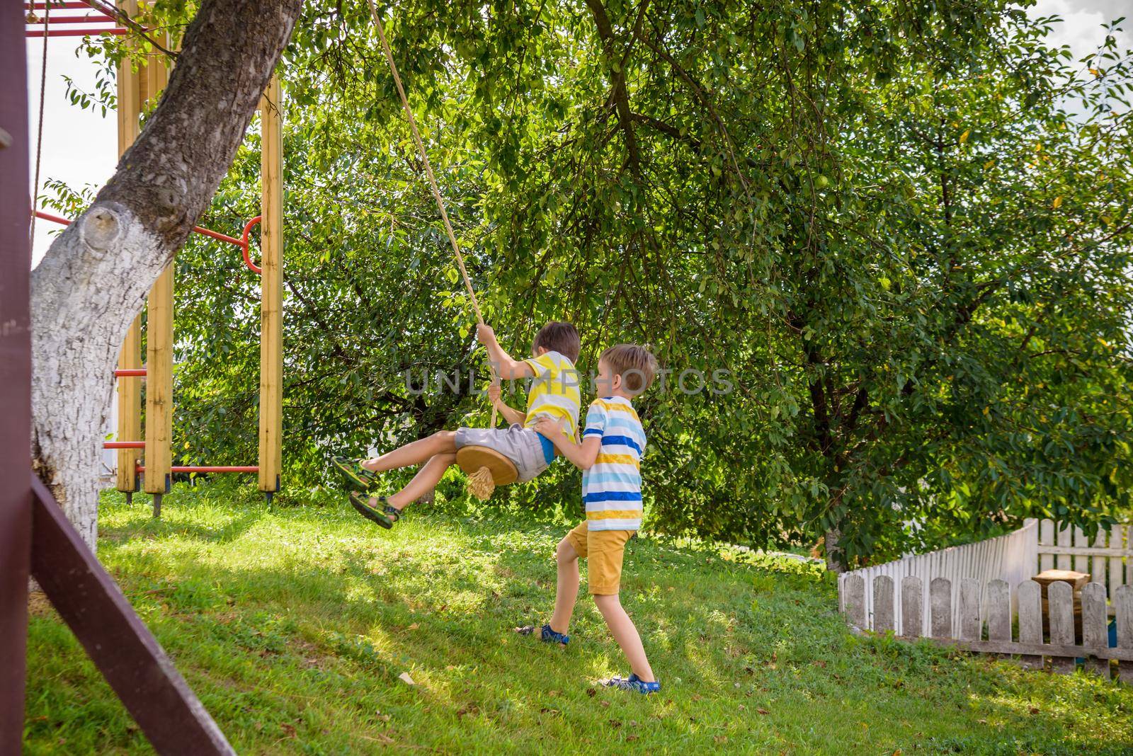 Two adorable happy little boys is having fun on a rope swing which he has found while having rest outside city. Active leisure time with children by Kobysh