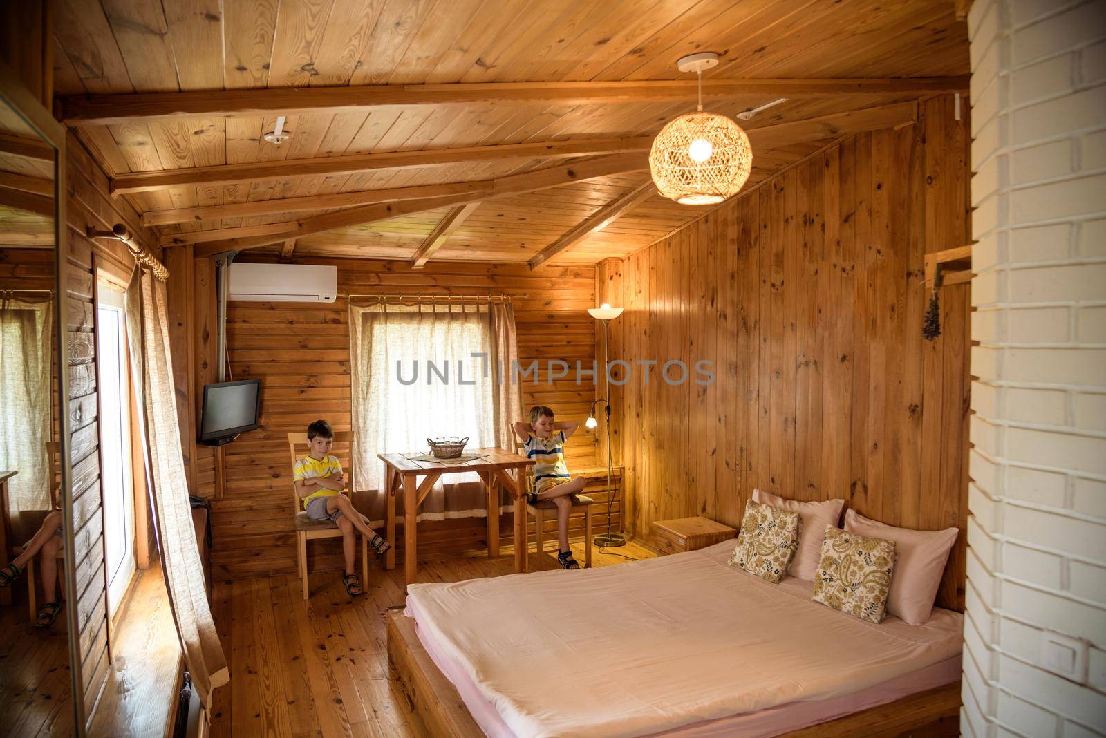 Two boys children in wooden living room in house with contemporary interior design, comfortable bad, carpet on floor, lamplight lamp, decor on table and wooden panel on copy space background.