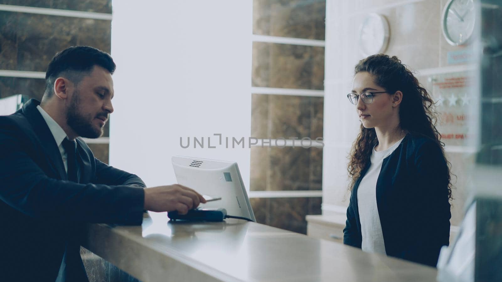 Businessman guest in hotel reception paying bill with smartphone via contactless payment technology and talking with receptionist standing at desk. Business, travel and people concept