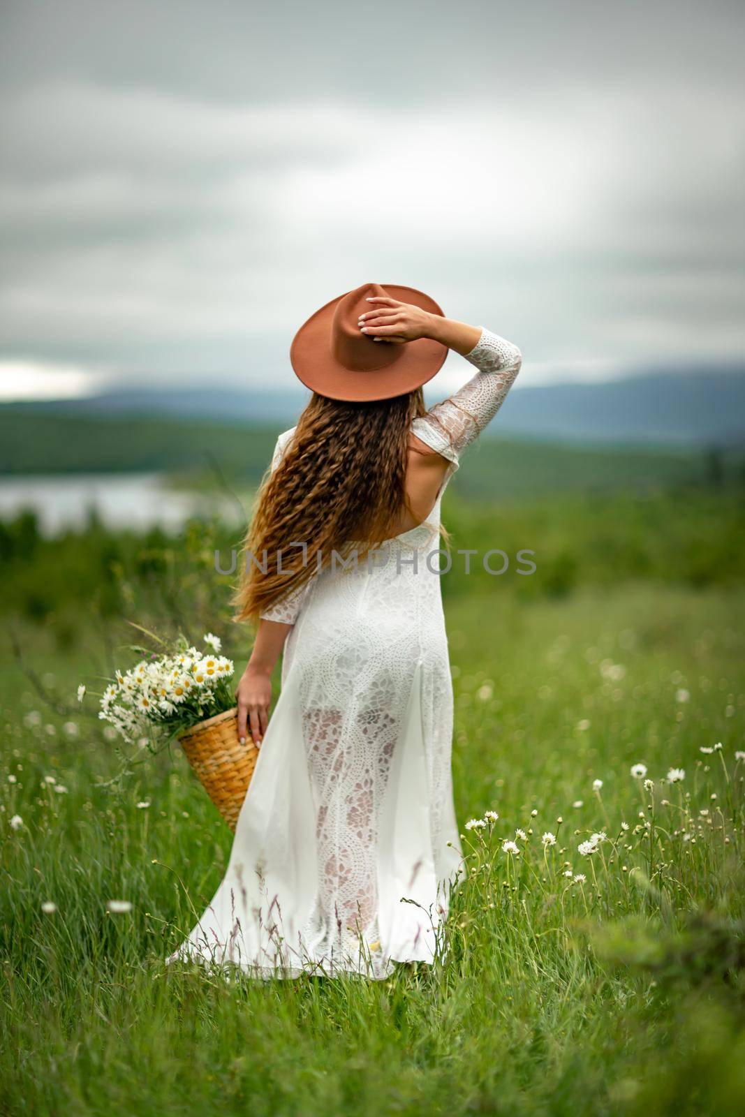 A middle-aged woman in a white dress and brown hat stands with her back on a green field and holds a basket in her hands with a large bouquet of daisies. In the background there are mountains and a lake. by Matiunina