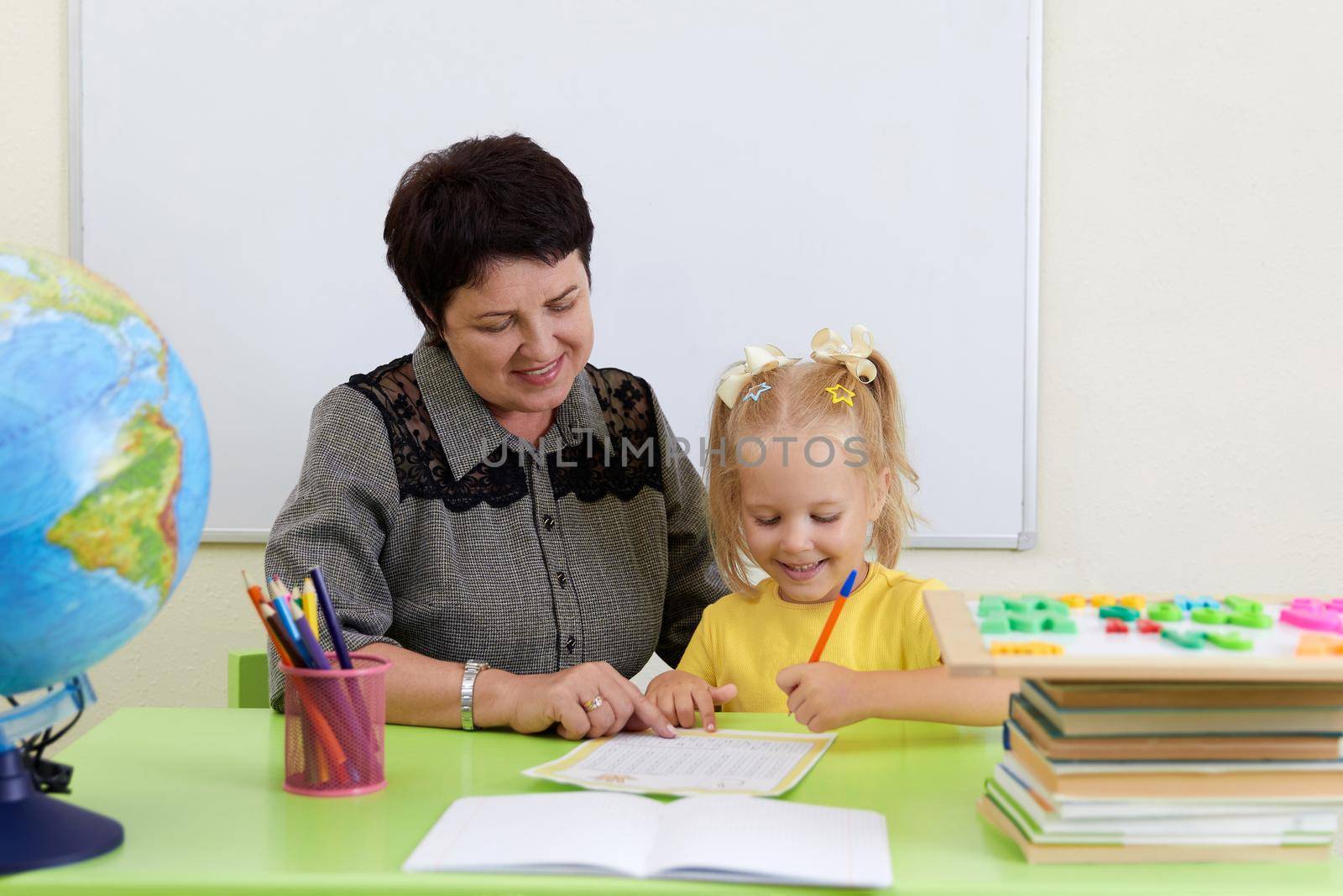 Mature teacher sitting at table teaching little girl how to write by Mariakray