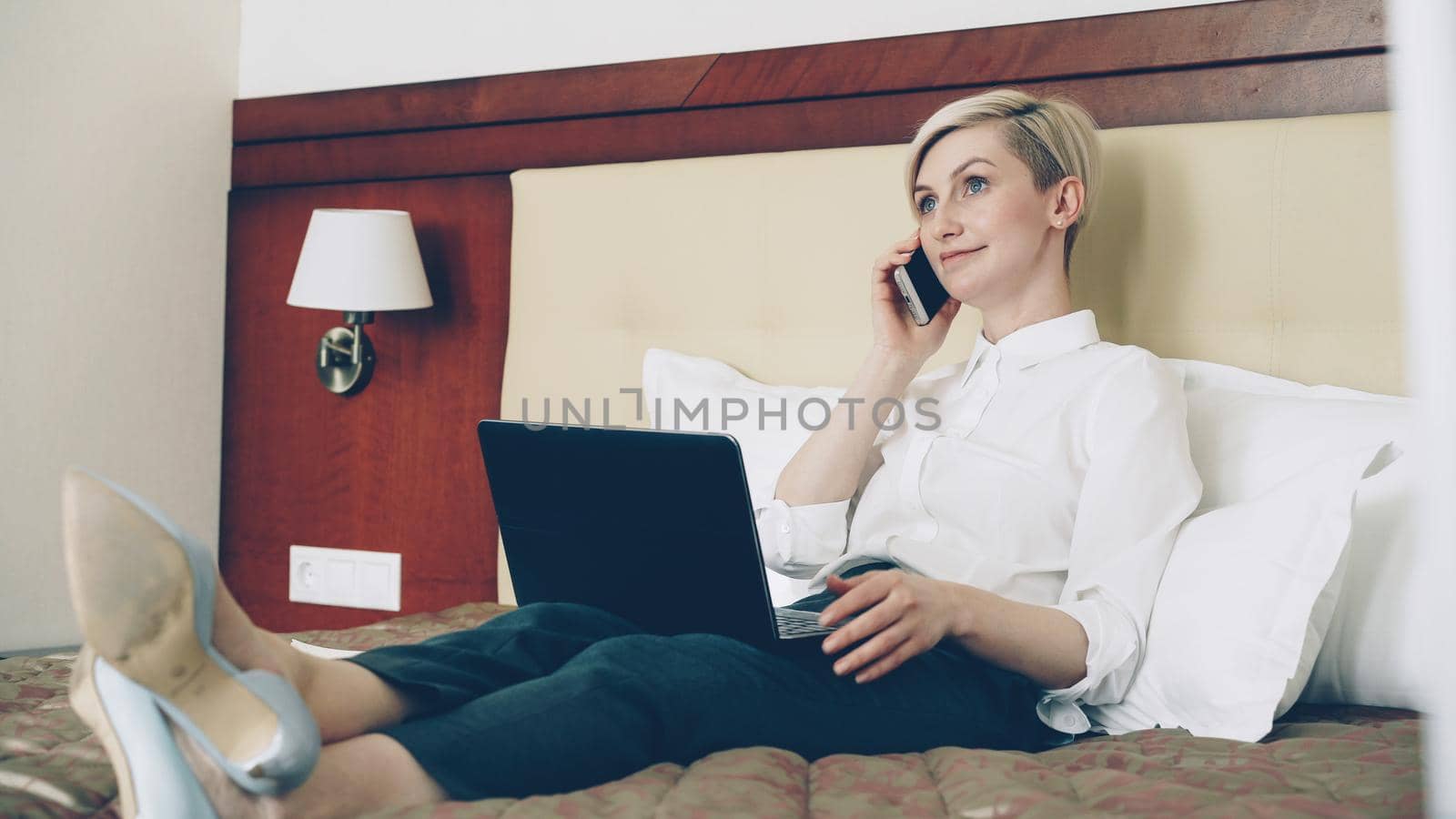 Smiling businesswoman in white shirt using on laptop and talking at mobile phone while lying on bed in hotel room. Business, travel and people concept by silverkblack