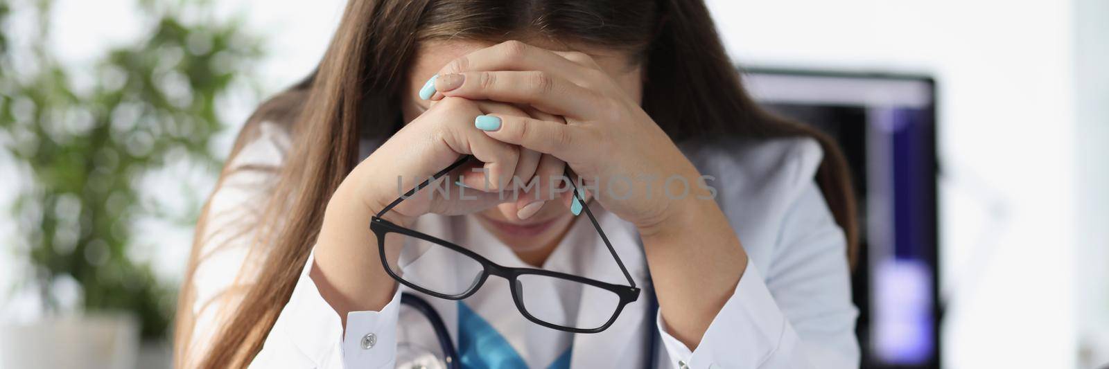 Tired woman doctor sitting at table and holding glasses in clinic. Treatment of critical patients concept