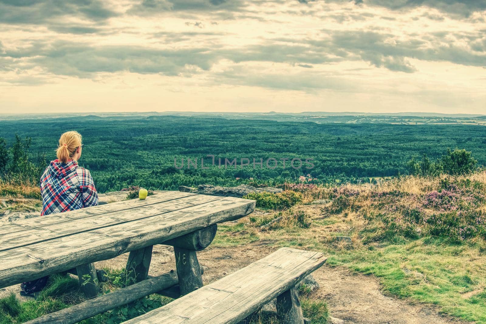 Young smiling blond girl trekker resting at wooden table with amazing view into landscape. Tired hiker sitting down and taking a break from hike. 
