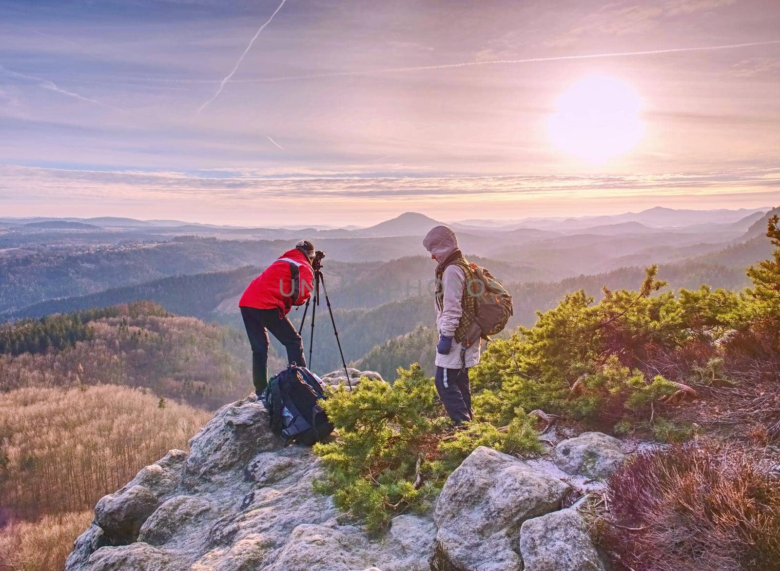 Hiker and photographer stay with tripod on cliff and takes photos. Two people stay at  tripod on the background of a mountain range and sky with clouds 