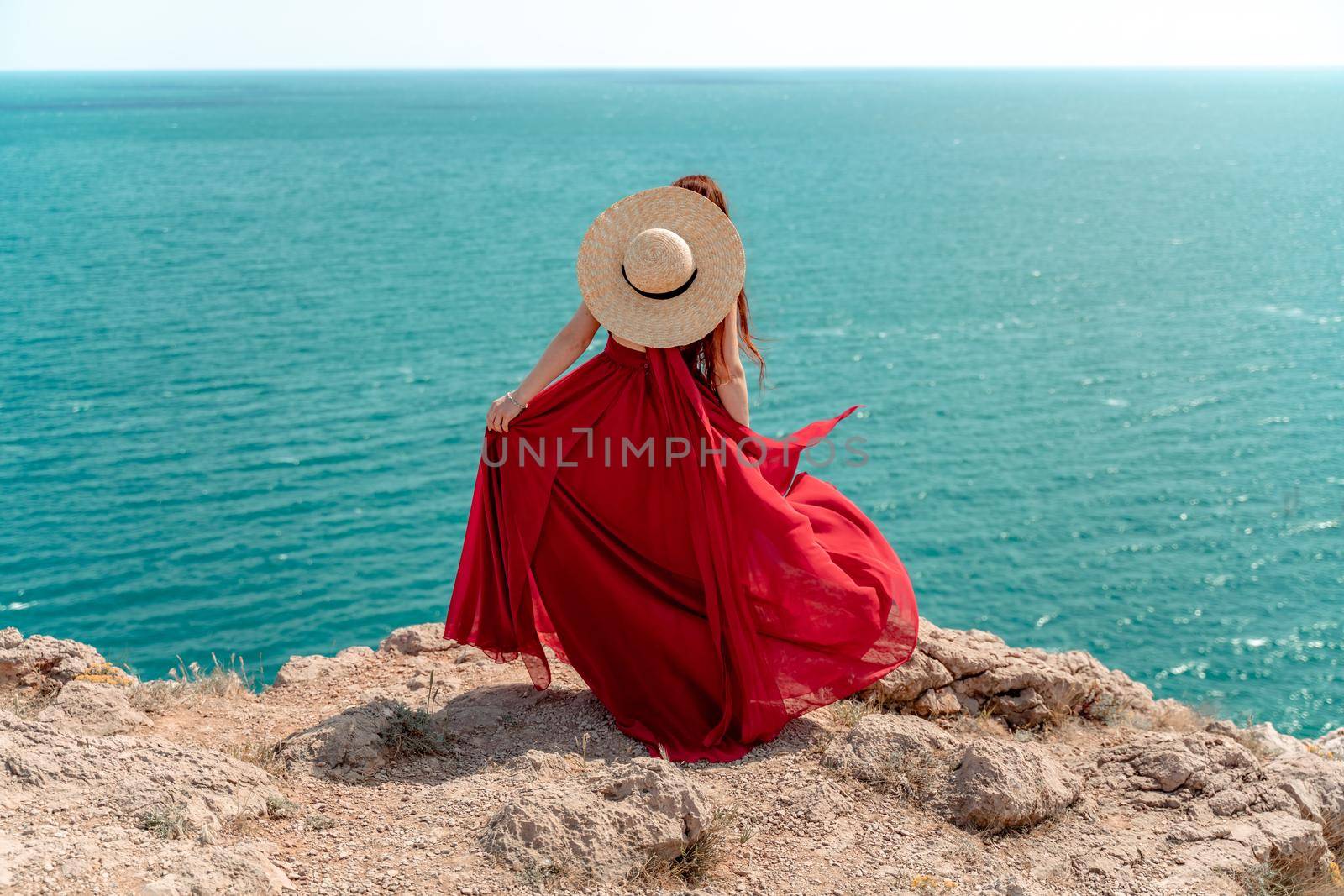 A woman, rear view in a red flying dress fluttering in the wind, a girl in a fluttering dress on the background of the sea. A straw hat hangs at the back of the neck. by Matiunina