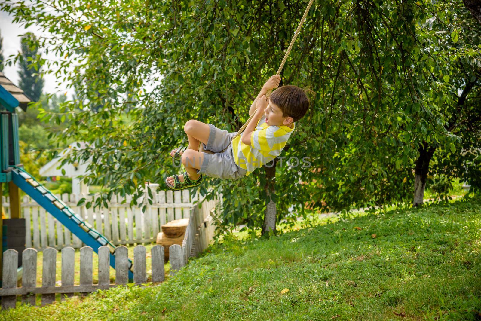 Happy little boy is having fun on a rope swing which he has found while having rest outside city. Active leisure time with children.