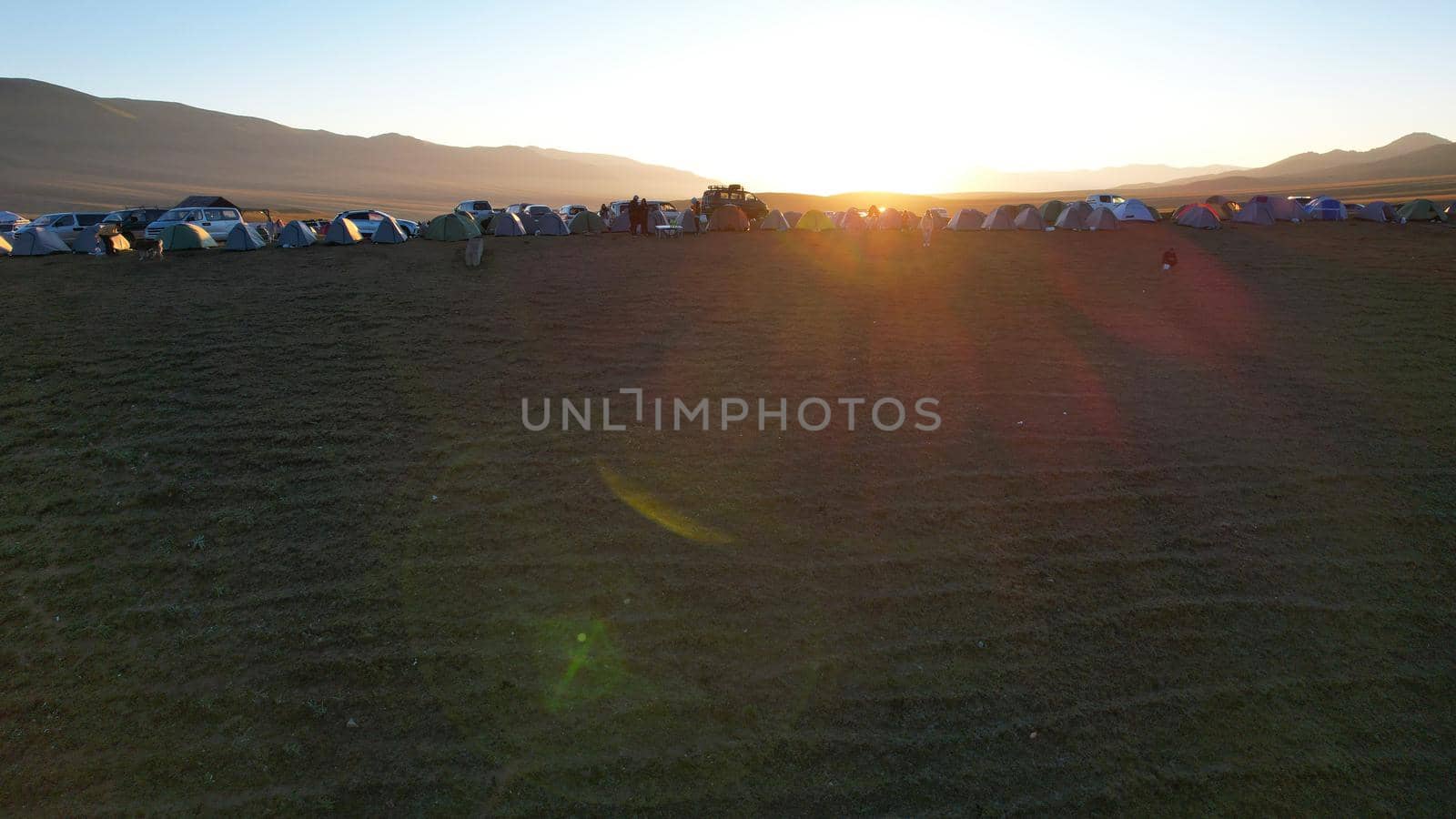 Drone view of a tent camp among green fields at dawn. There are cars nearby, tents of different colors. People are just waking up. Clear sky. Mountains and rays of the sun are in the distance. Plain