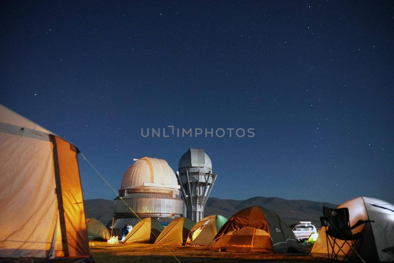 A camp with colored tents near the observatory. The Perseid starfall. A large teleskom watches the starry night sky. Meteorites and planes fly by, leaving traces in the sky. The stars are burning.