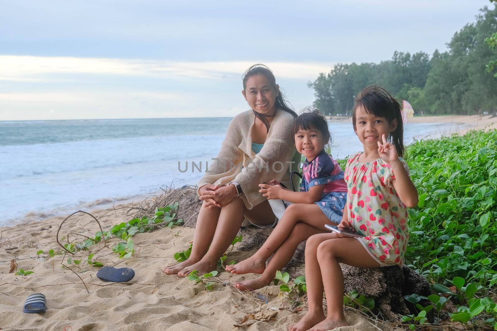 Happy Asian family of mother and daughters sitting on a log smiling at the camera on the beach during a summer vacation at sunset. Summer family trip to the beach. travel and vacation concept.