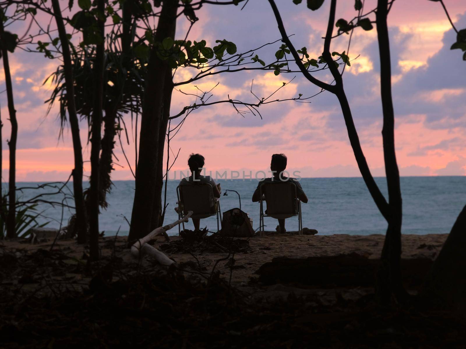 Couples sit and watch the sunset by the sea. Silhouettes of women and men sitting on a bench watching the serene sunset over the ocean. Relaxation and vacation travel concept