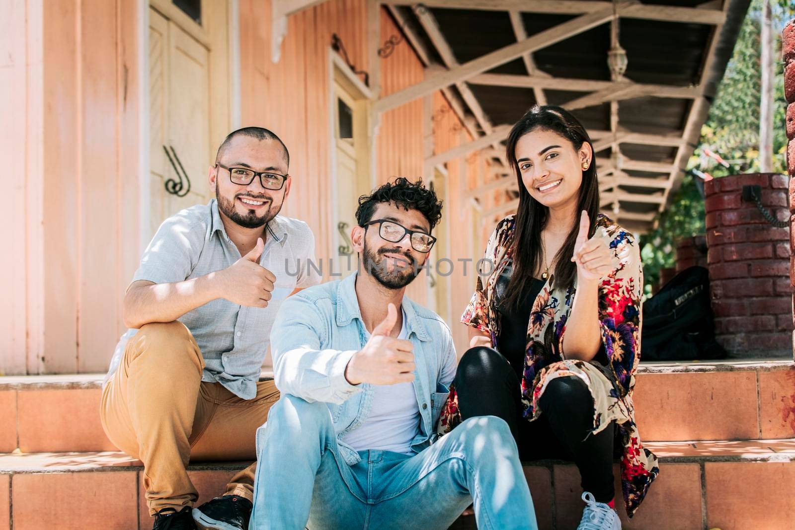 Group of happy friends sitting on the stairs giving thumbs up. Portrait of happy teenagers sitting outside giving thumbs up, Close up of happy sitting students giving thumbs up by isaiphoto