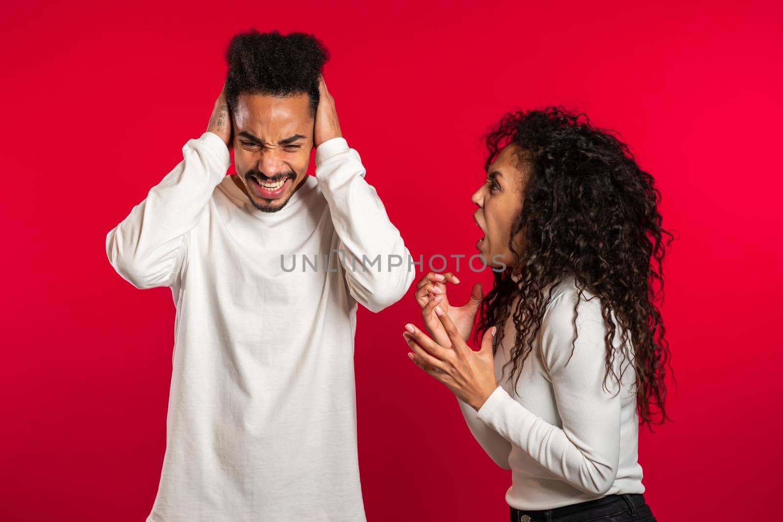 Young african woman emotionally screaming at her husband or boyfriend on red background in studio. Bored man covers ears with hands. Concept of conflict, problems in relationships