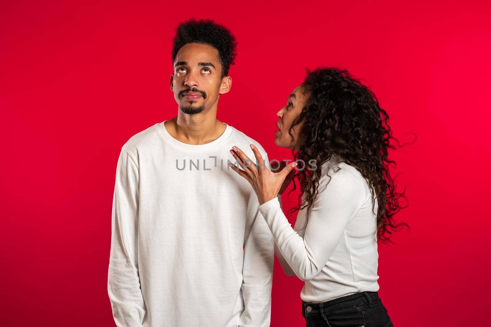 Young african woman emotionally screaming at her husband or boyfriend on red background in studio. Bored man rolling his eyes. Concept of conflict, problems in relationships