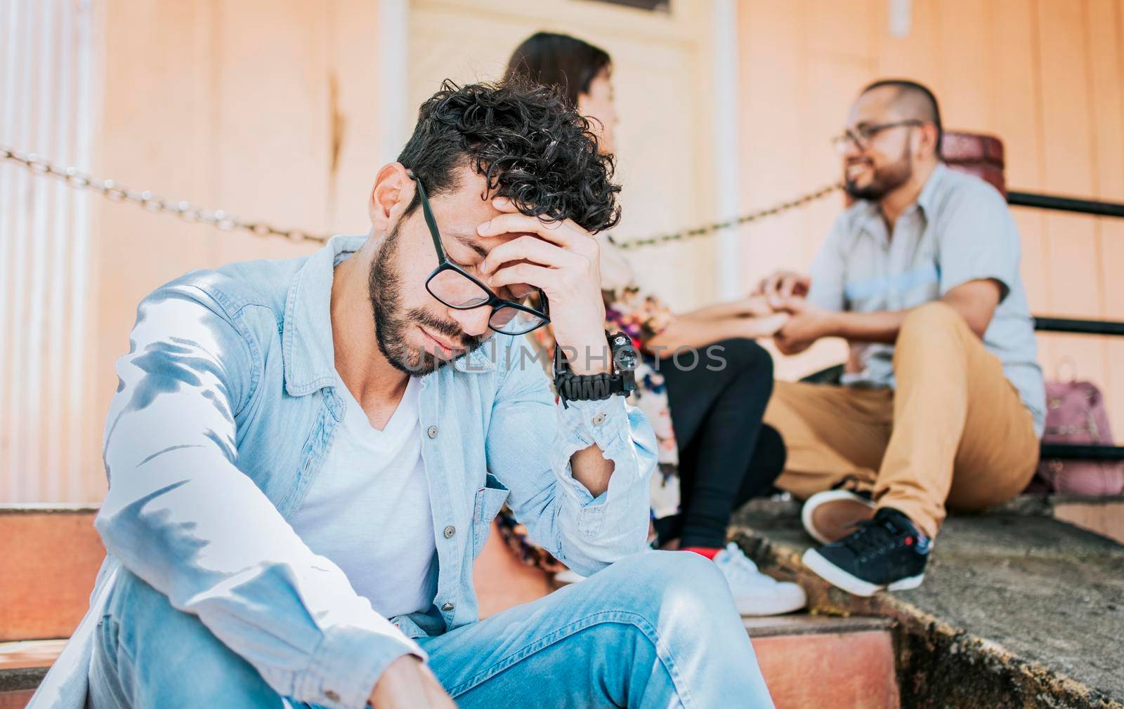 Sad boyfriend sitting on some stairs while his girlfriend talking to another man. Sad man seeing his girlfriend cheating with his friend. Girl talking to another man and her boyfriend is offended by isaiphoto