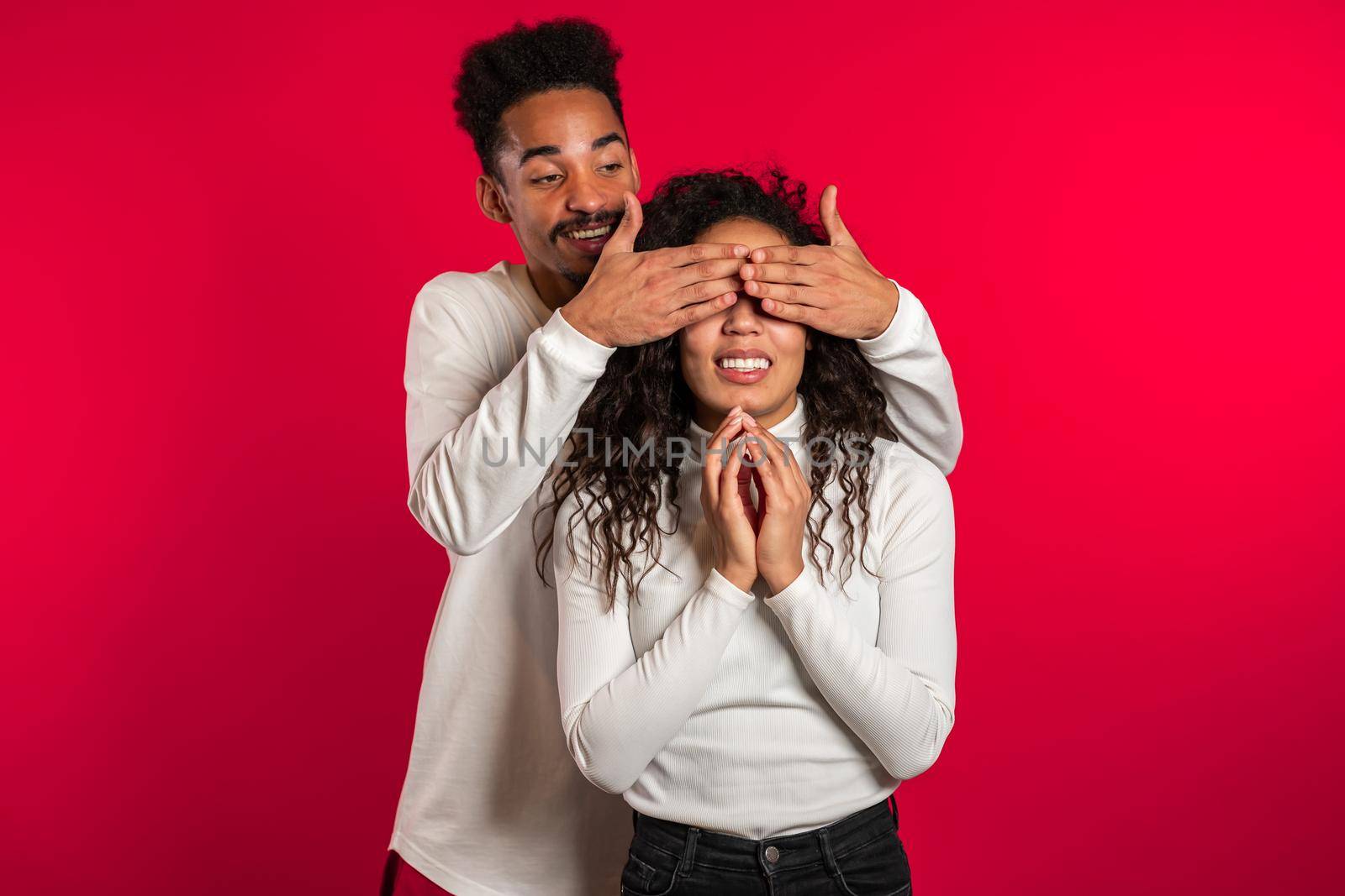 African man closes eyes of his beloved girlfriend before surprise her. Couple in white on red studio background. Love, holiday, happiness concept