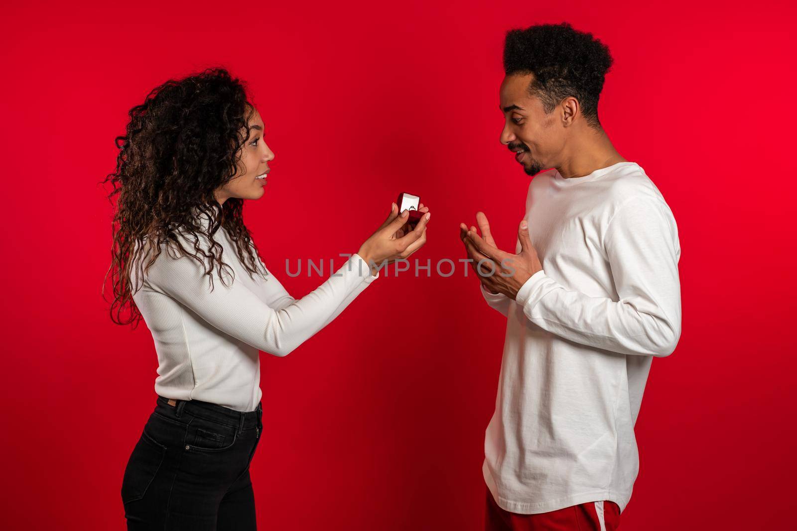 Young couple. African woman makes marriage proposal to her lover man with ring on red studio background. Feminism, equality rights concept.