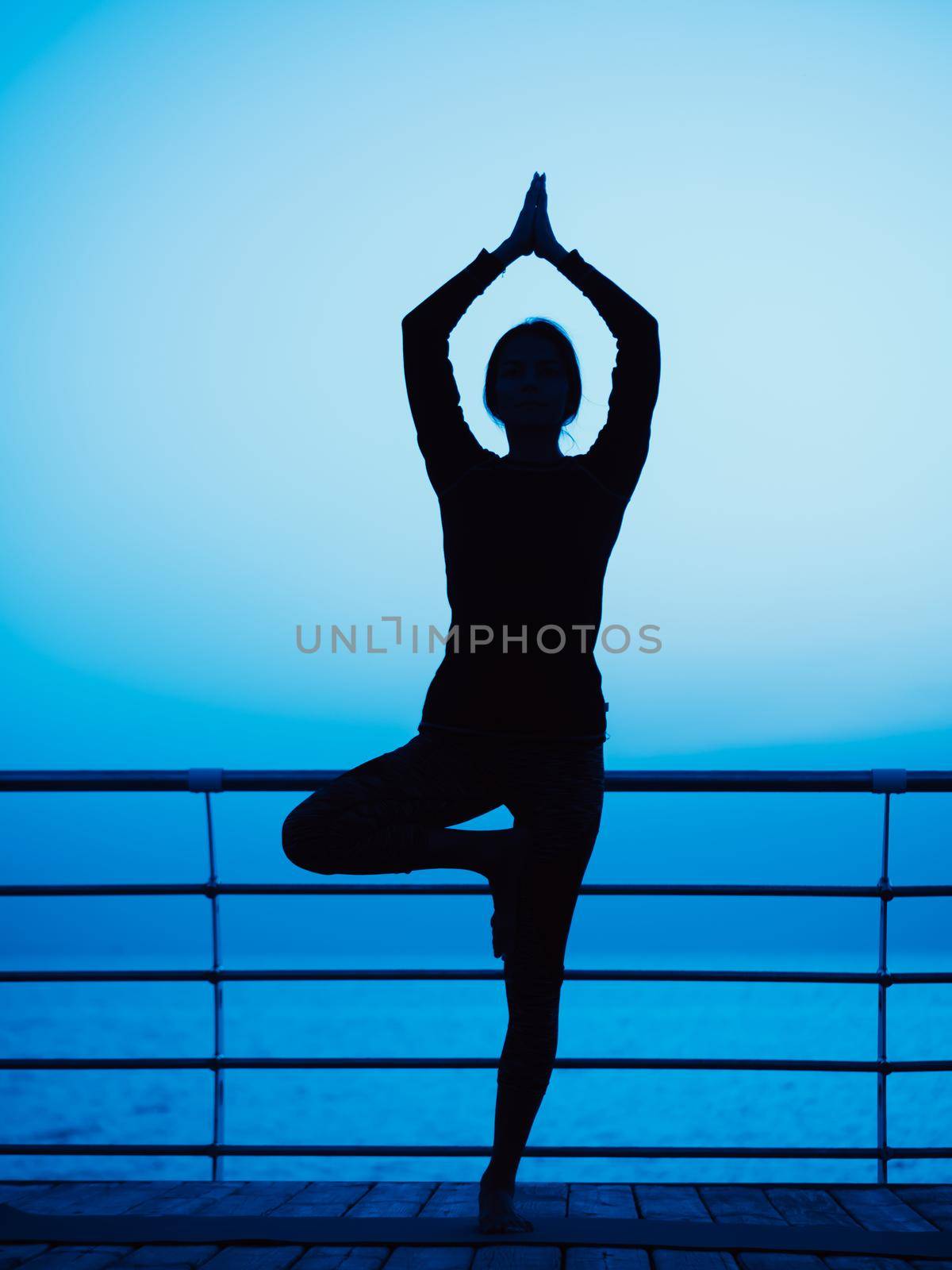 Young slim girl practicing yoga on embankment against ocean or sea at night time. Silhouette of woman in asana of tree.