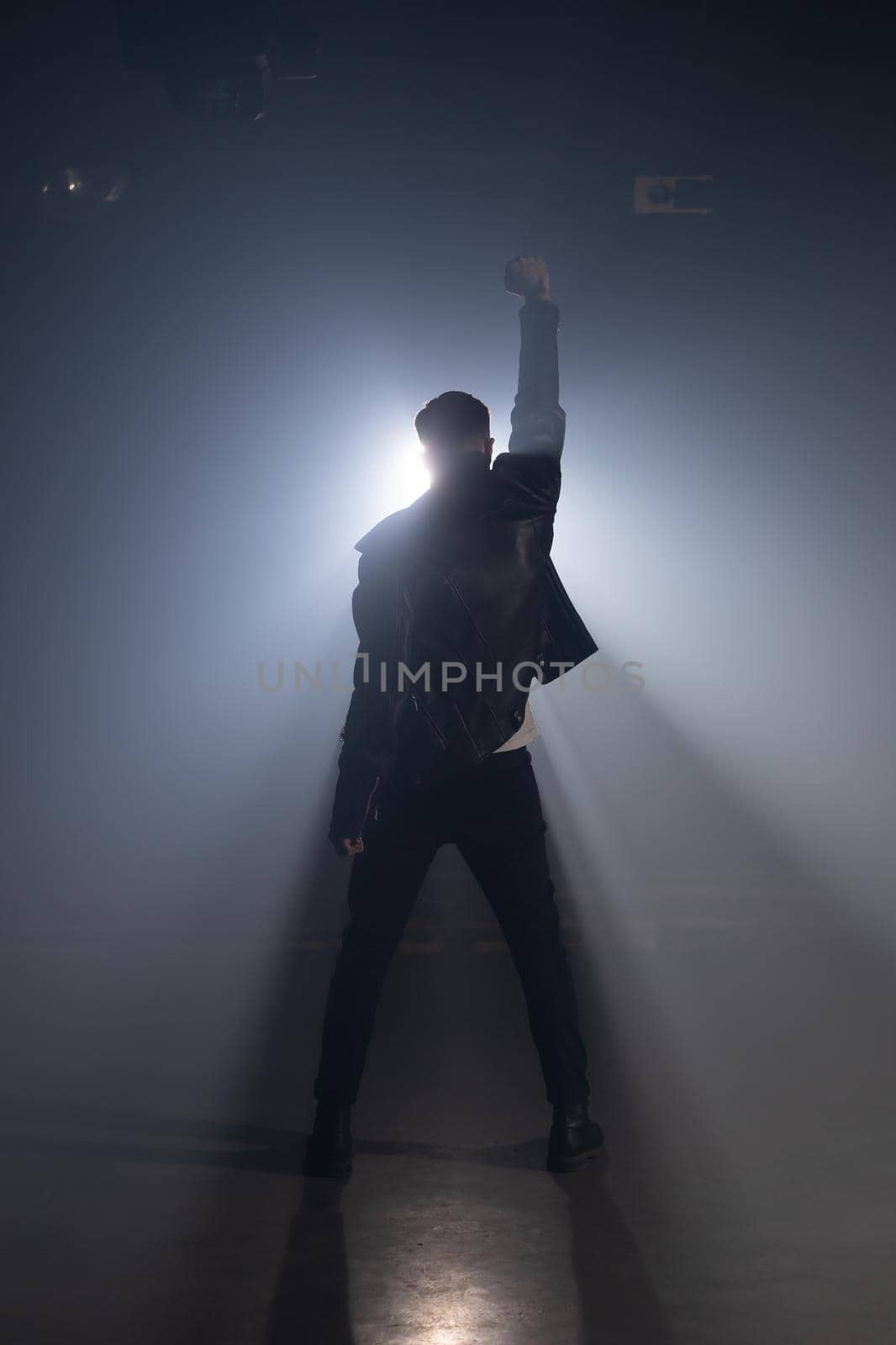 Young man in leather jacket on smoky stage. View of guy raises hand up as gesture and symbol of success, freedom and victory