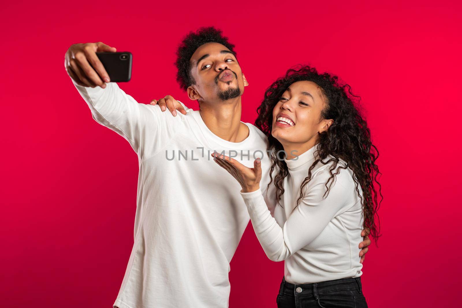 Young african american couple in white making selfie with smartphone on red studio background.Lovers match, love, holidays, happiness concept.