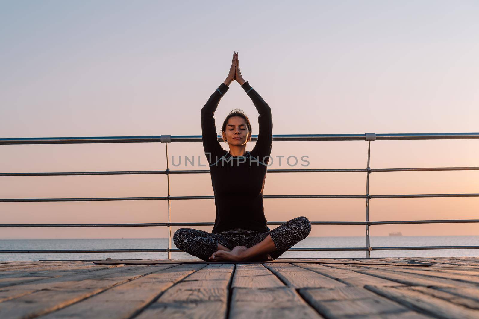 Concentrated girl in black practicing in yoga meditation on wooden seafront. Woman in lotus pose. Stretching, practice, healthy lifestyle concept. Copy space by kristina_kokhanova