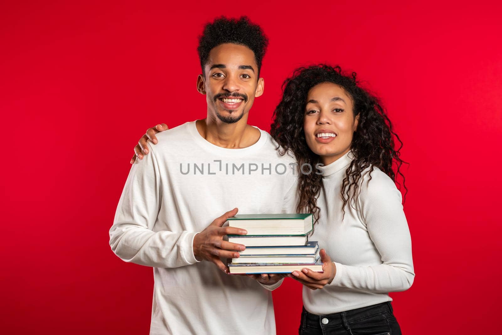 African students on red background in studio holds stack of university books from library. Classmates smiles, they happy to graduate