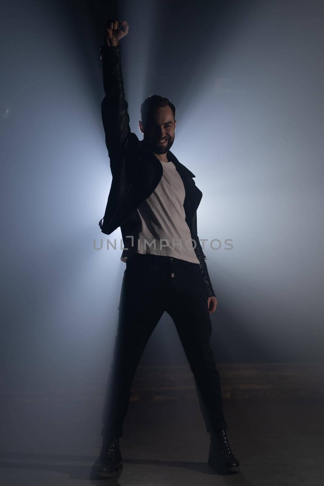 Young man in leather jacket on smoky stage. View of guy raises hand up as gesture and symbol of success, freedom and victory