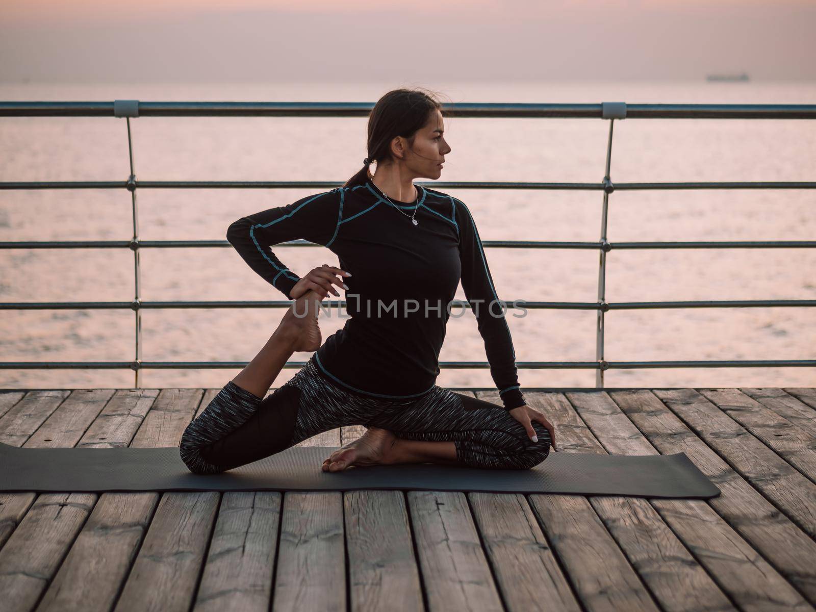 Young brunette woman doing yoga exercises on wooden sea embankment in the morning. Girl in black sports costume. Stretching, practice, healthy lifestyle concept.