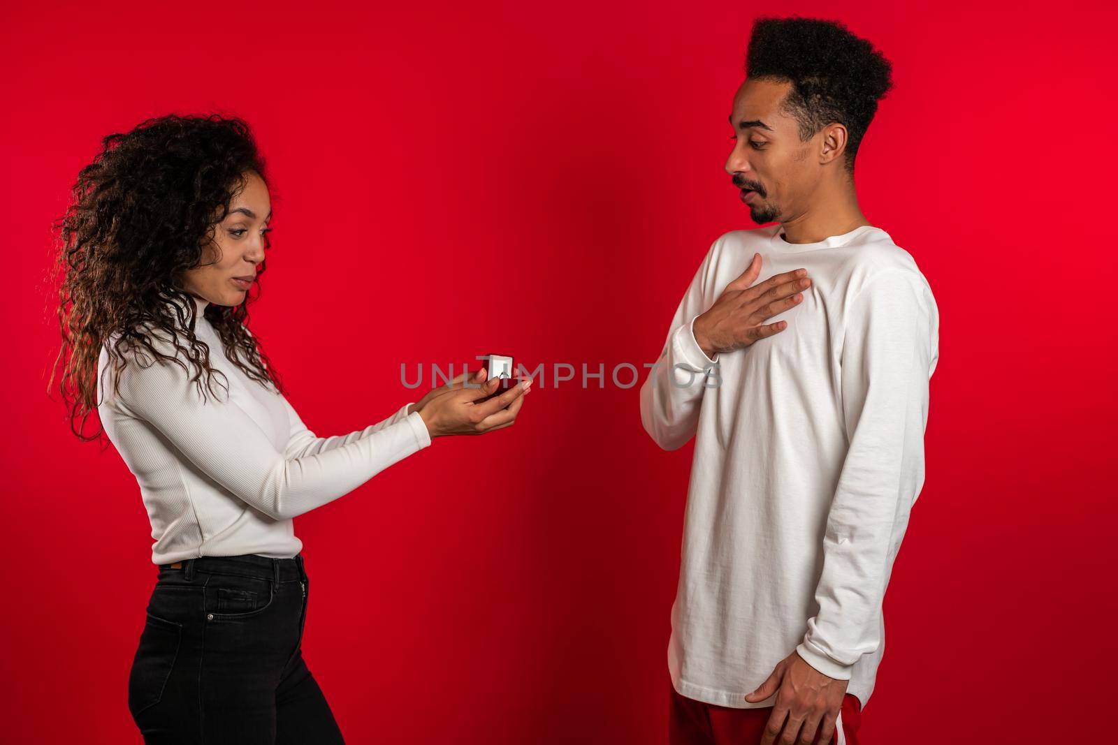 Young couple. African woman makes marriage proposal to her lover man with ring on red studio background. Feminism, equality rights concept.