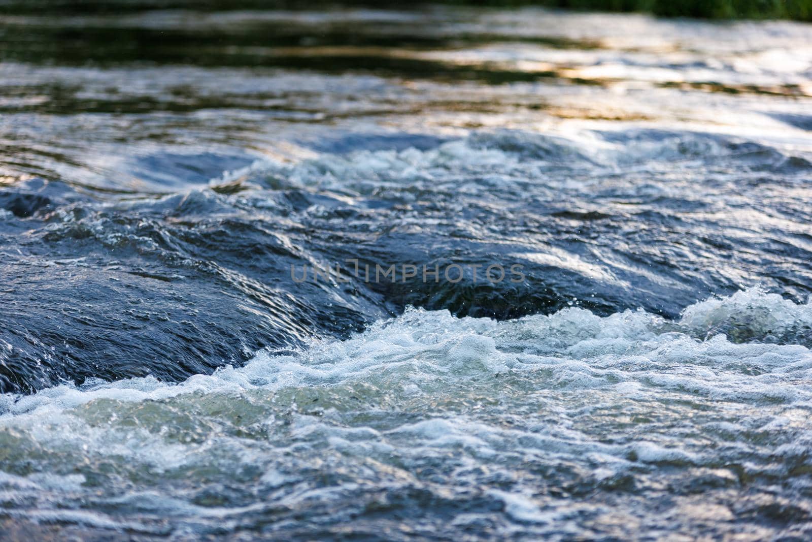 flowing water of a summer river with a small rapid waterfall at evening light, short or fast shutter speed, one 400-th sec exposure