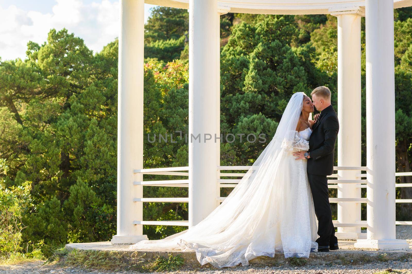 Portrait of mixed-racial newlyweds against the background of a gazebo with round columns, a girl in a lush white dress by Madhourse