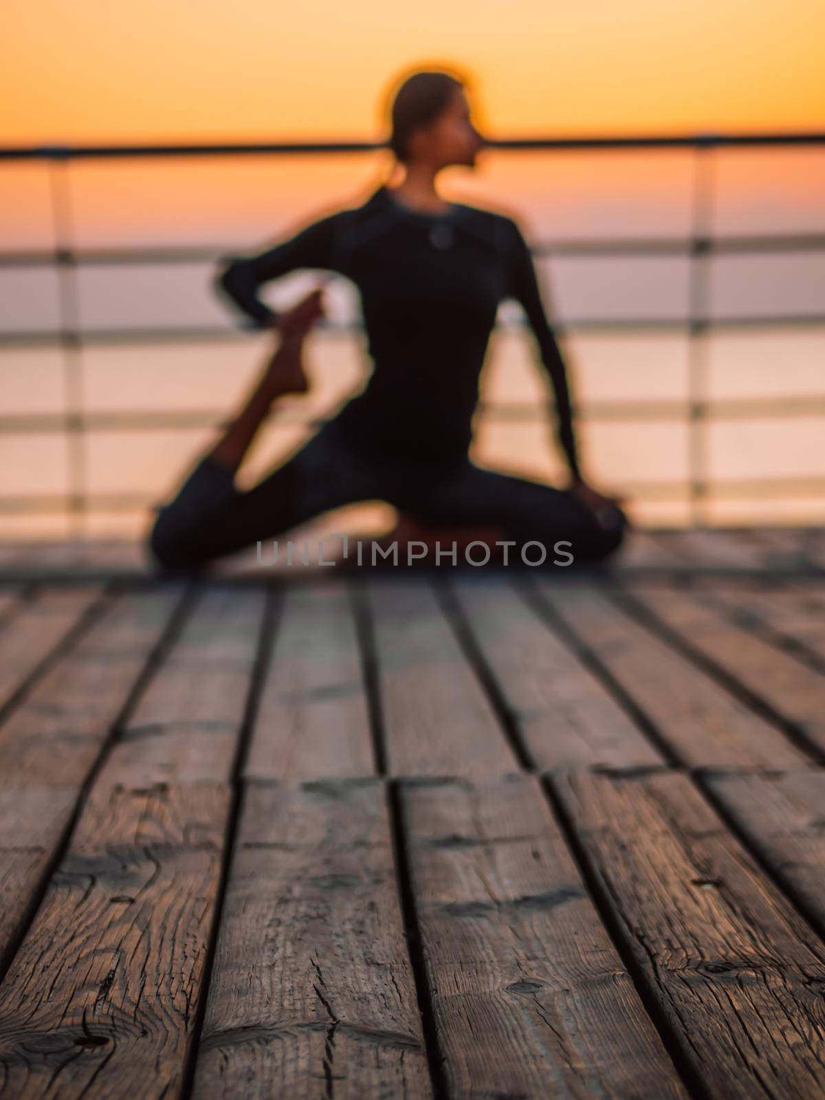 Young blurred woman doing yoga exercises on wooden sea embankment in the morning. Girl in black sports costume. Stretching, practice, healthy lifestyle concept.