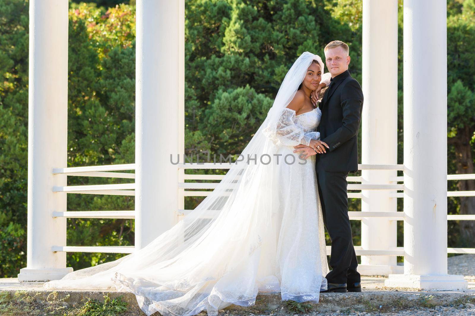 Portrait of a mixed race newlyweds in front of a gazebo with round columns by Madhourse