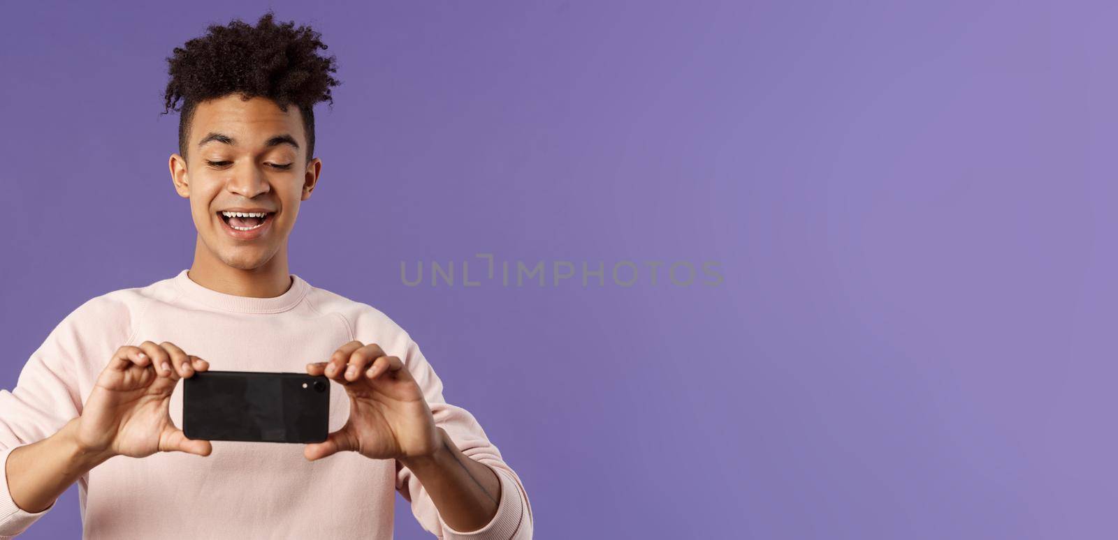 Portrait of amazed, excited young man seeing something interesting, stream concert to his internet social network profile, taking photo or recording video with mobile phone, purple background.