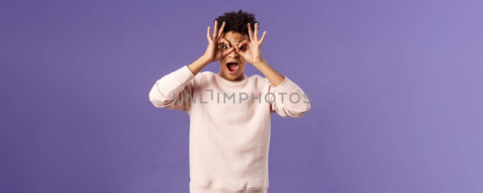 Portrait of funny, enthusiastic young hispanic man fooling around, making finger glasses, mask with hands over eyes, open mouth fascinated and surprised, seeing great product or promo action by Benzoix