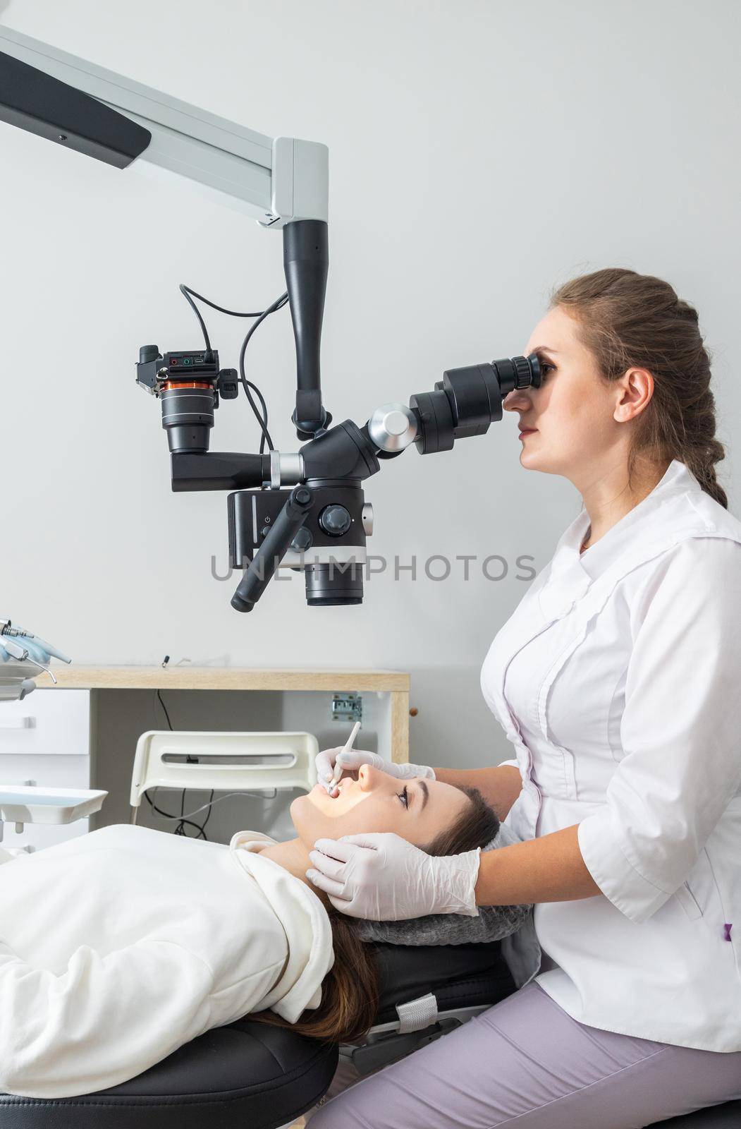 Female dentist using dental microscope treating patient teeth at dental clinic office. Medicine, dentistry and health care concept. Dental equipment by Mariakray