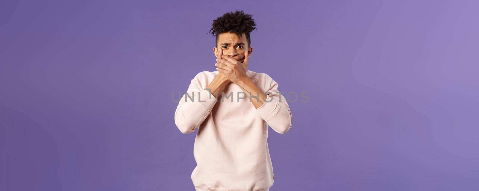 Shocked worried, embarrassed young man said something he shouldnt have, shut his mouth with hands and look guilty or anxious at camera, feel sorry for being rude, purple background.