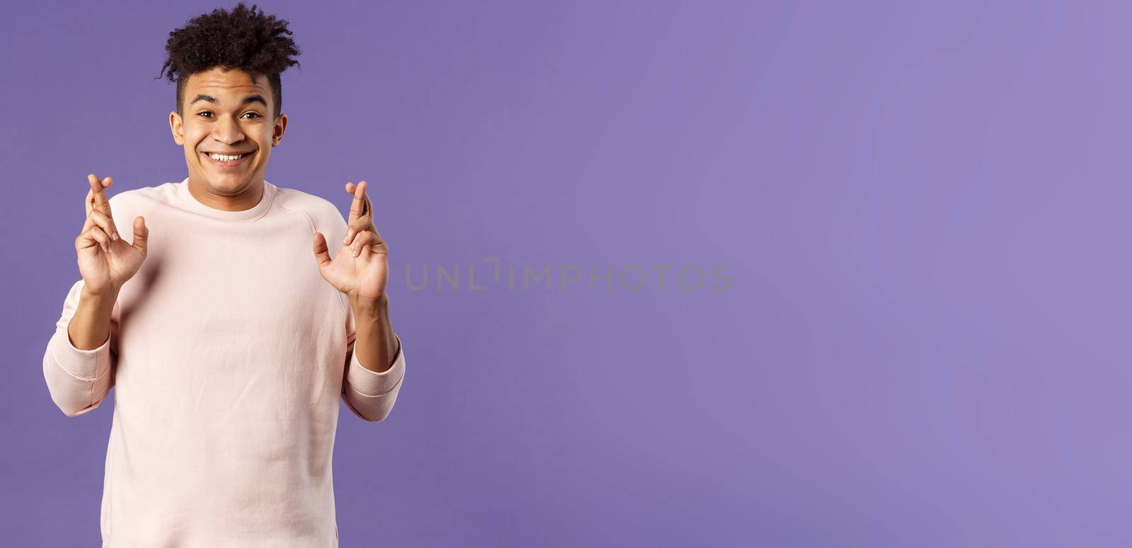 Portrait of hopeful positive, smiling young man having faith in dreams may come true, make wish, cross fingers good luck and awaiting for miracle, standing purple background praying.