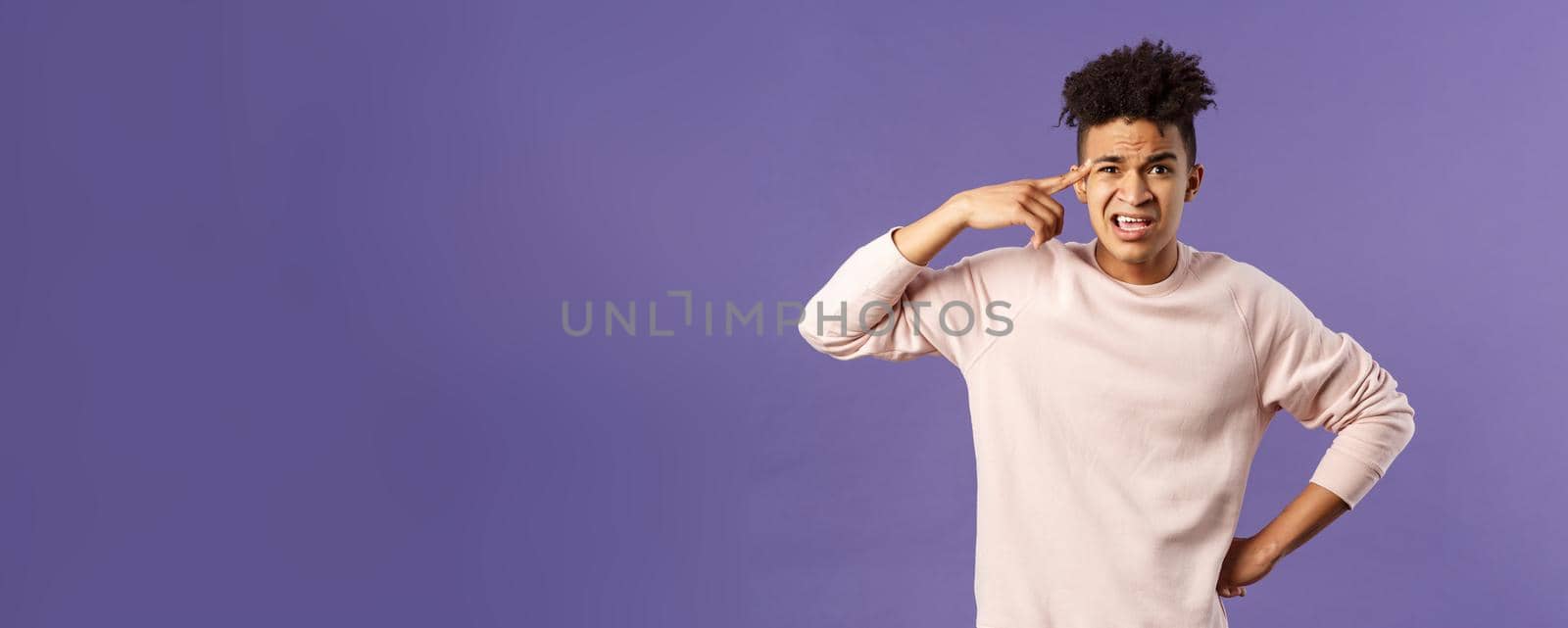 Portrait of angry, annoyed young man scolding someone from being stupid and crazy, rolling index finger over temple staring outraged and irritated camera, standing bothered purple background.