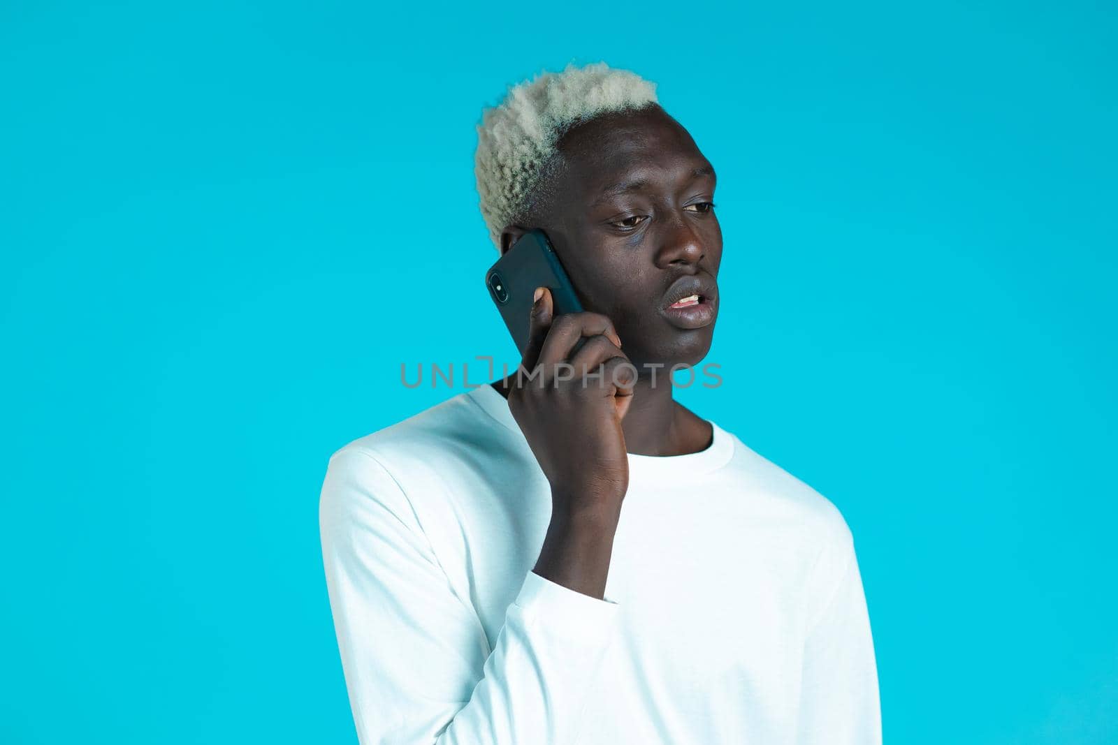 Young afro man speaks serious on phone. Guy holding and using smart phone. Blue studio background. by kristina_kokhanova