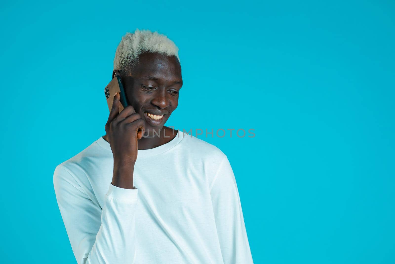 Young african american man speaks with smile on phone. Guy holding and using smart phone. Blue studio background. by kristina_kokhanova