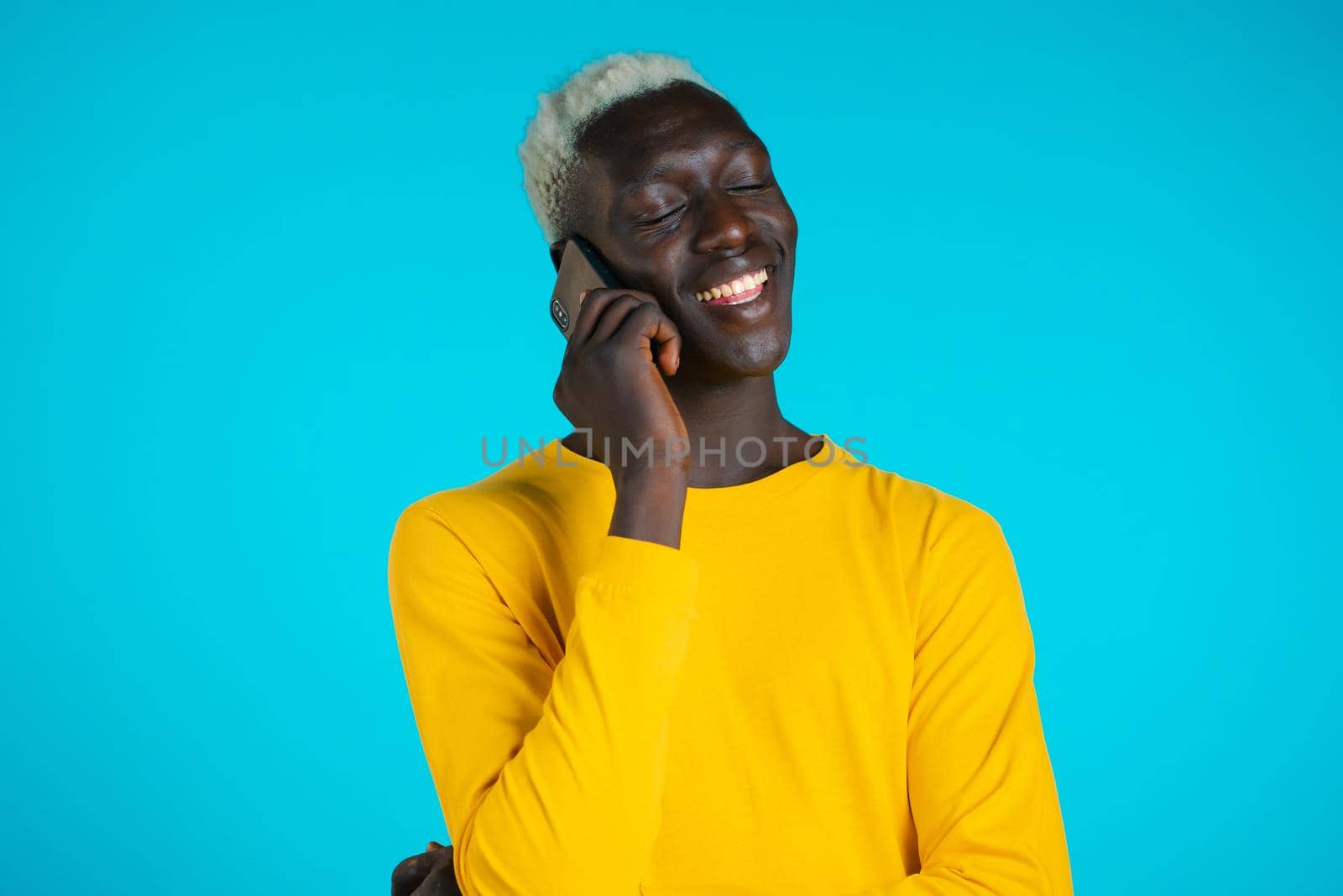 Young african american man speaks with smile on phone. Guy holding and using smart phone. Blue studio background