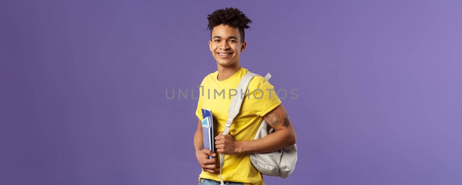 Back to school, university concept. Portrait of handsome charismatic hispanic guy, student walking to campus, going for lecture, studying hold backpack and notebooks, stand purple background by Benzoix