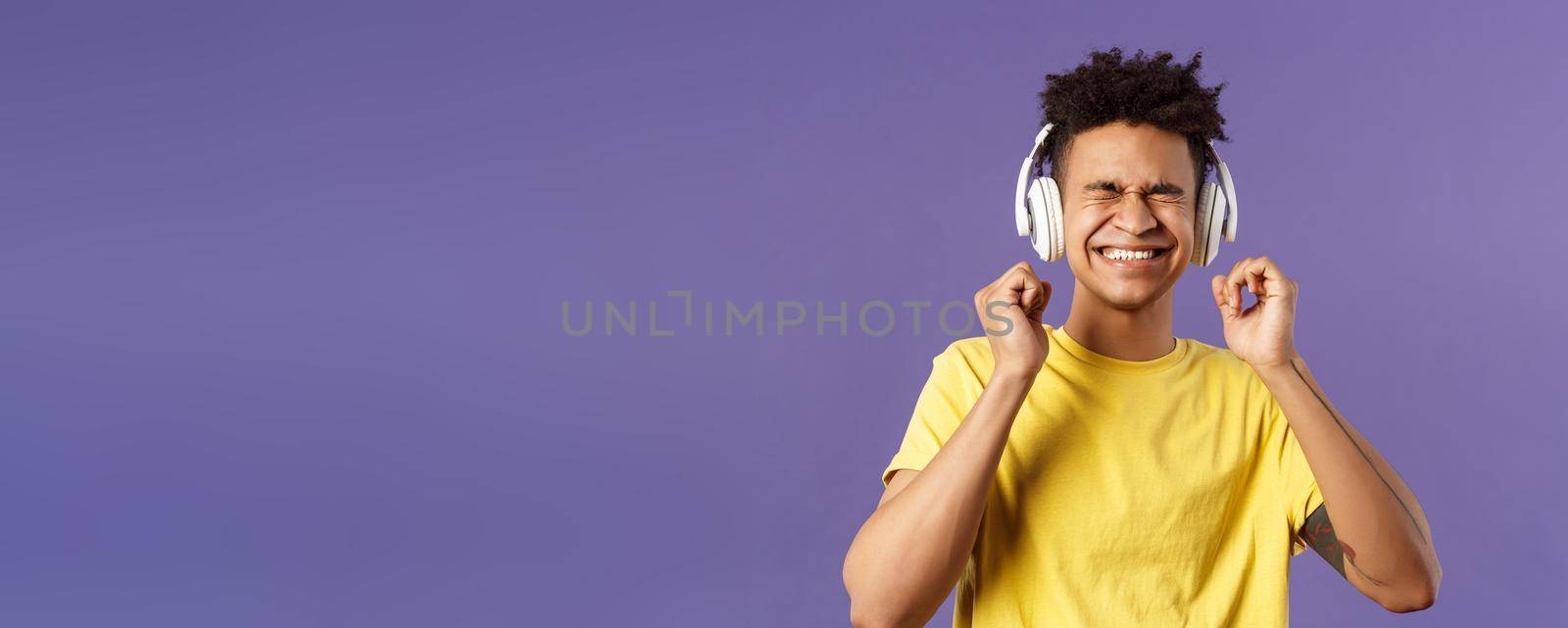 Portrait of pleased, excited young man enjoying nice quality awesome beats in headphones, close eyes and smiling rejoicing, dancing over cool new song, listen music over purple background.