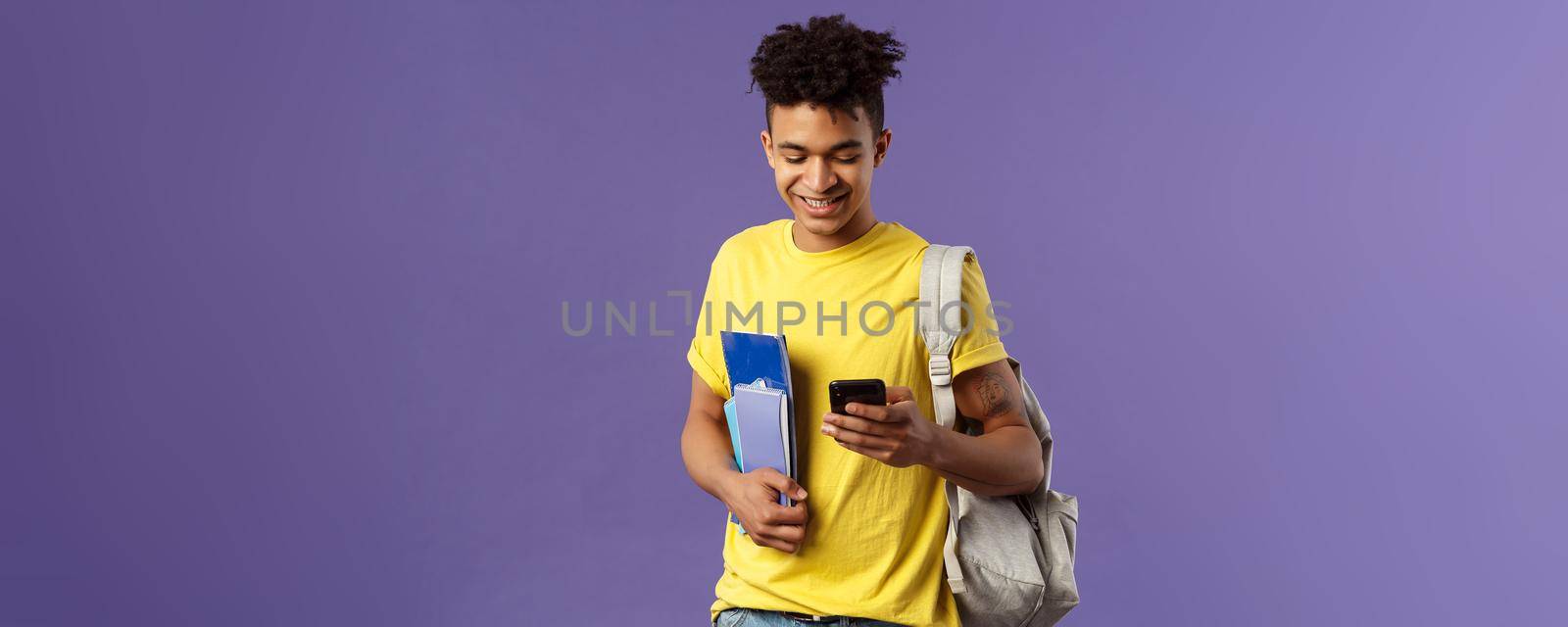 Back to school, university concept. Portrait of pleased good-looking male student texting friend, looking at mobile phone on his way to classes in campus, hold notebooks and backpack by Benzoix