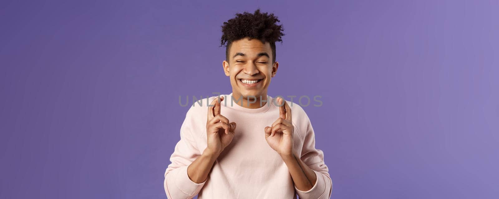 Close-up portrait of happy young hopeful guy anticipating something good happen, cross fingers good luck, close eyes and smiling awaiting miracle, praying for dream come true, purple background.
