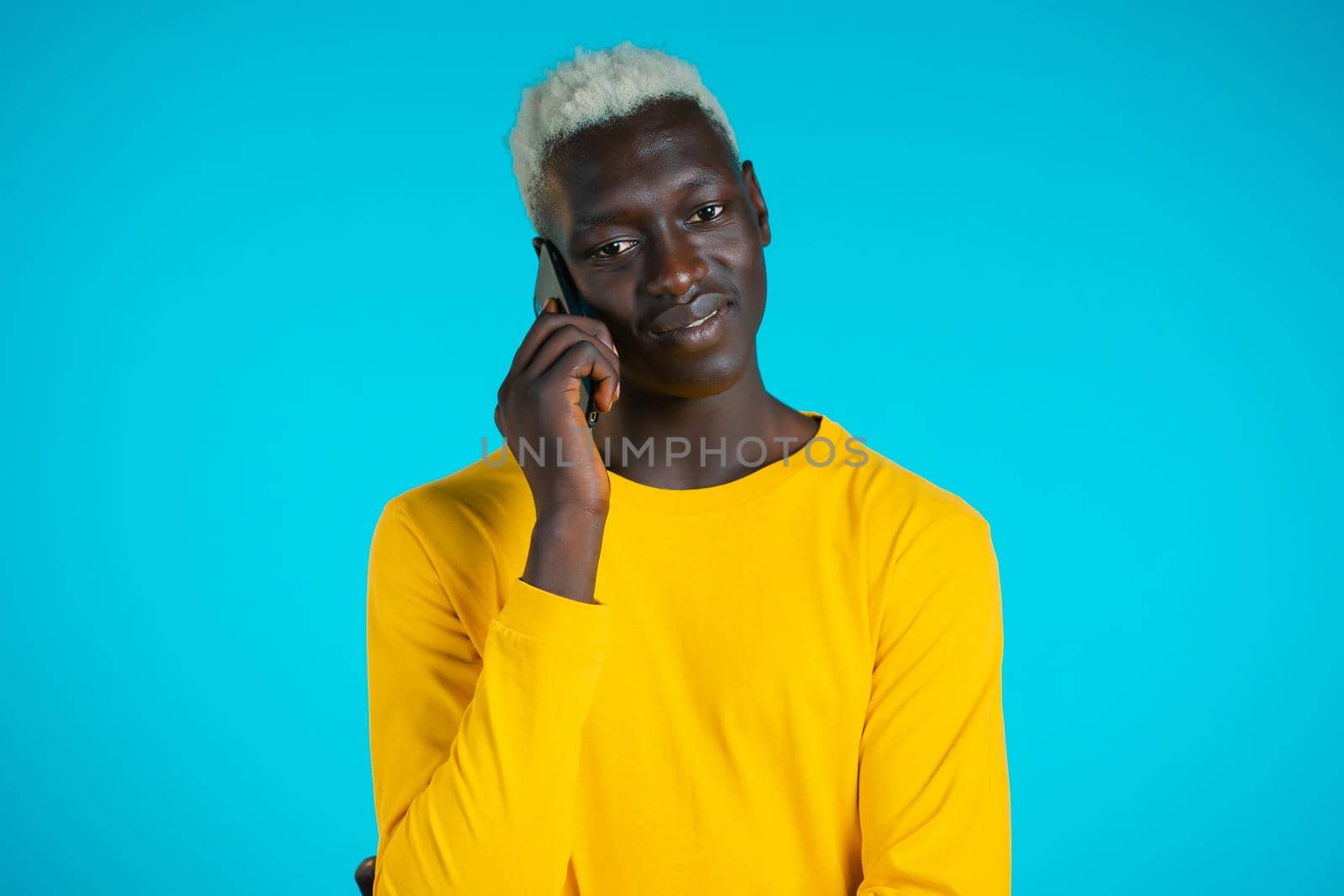 Young african american man speaks with smile on phone. Guy holding and using smart phone. Blue studio background