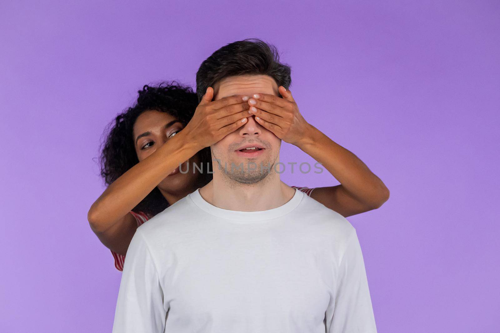 African american woman closes eyes of her beloved boyfriend before surprise him. interracial couple on purple studio background. Love, holiday, happiness concept. High quality photo